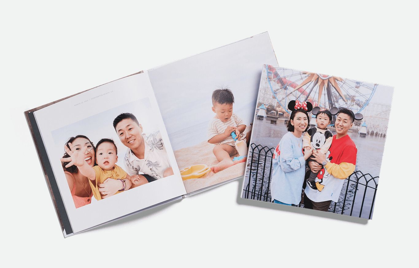 A stack of photo books stands upright, with the front book displaying a baby in a sun hat sitting on grass next to a dog in a red hat, both in front of a wooden fence.