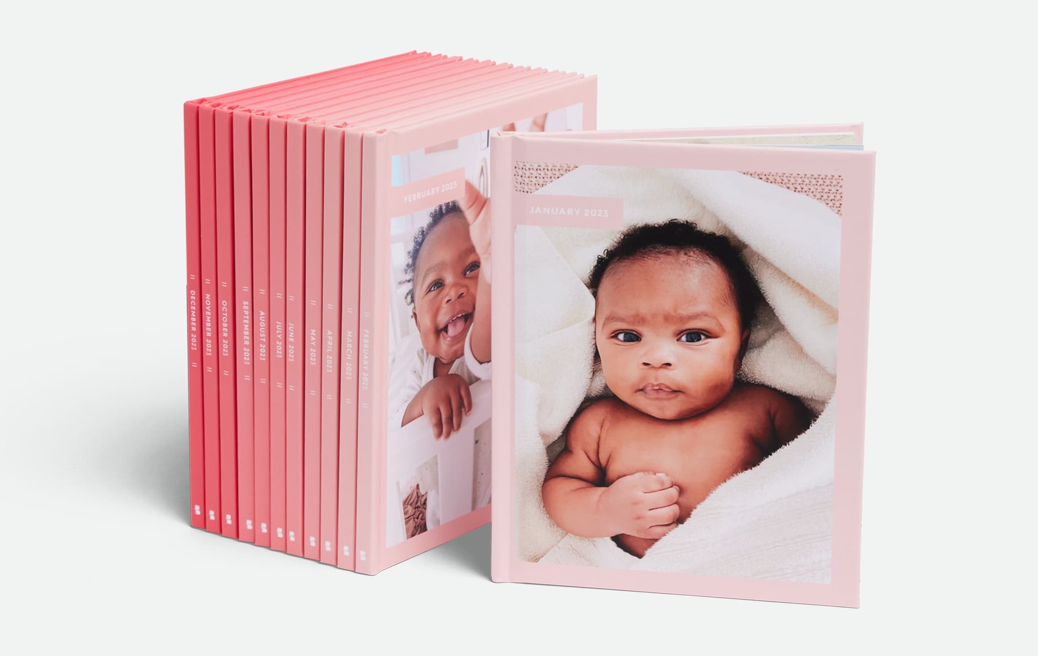 A stack of photo books stands upright, with the front book displaying a baby in a sun hat sitting on grass next to a dog in a red hat, both in front of a wooden fence.