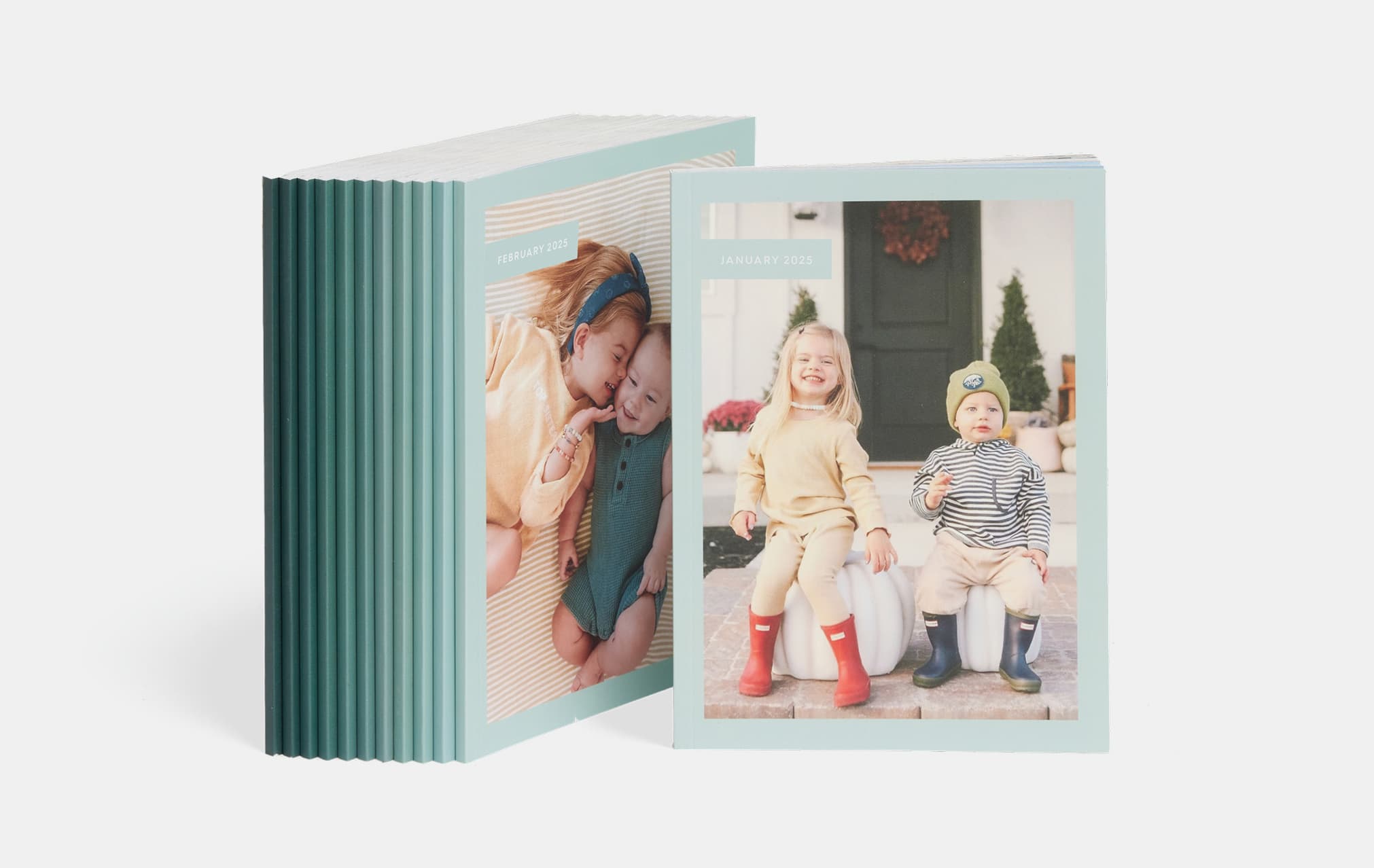 A stack of photo books stands upright, with the front book displaying a baby in a sun hat sitting on grass next to a dog in a red hat, both in front of a wooden fence.