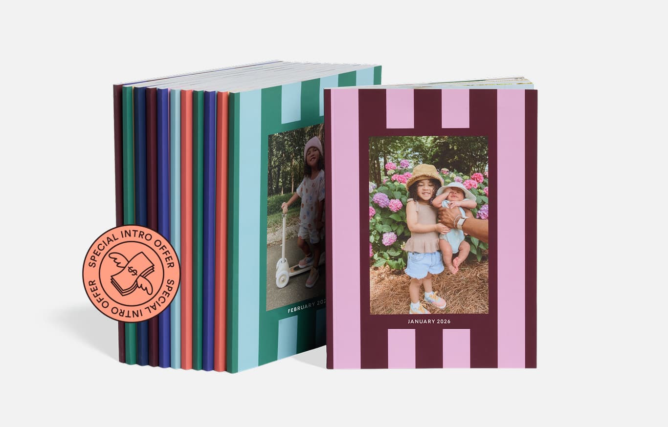 A stack of photo books stands upright, with the front book displaying a baby in a sun hat sitting on grass next to a dog in a red hat, both in front of a wooden fence.