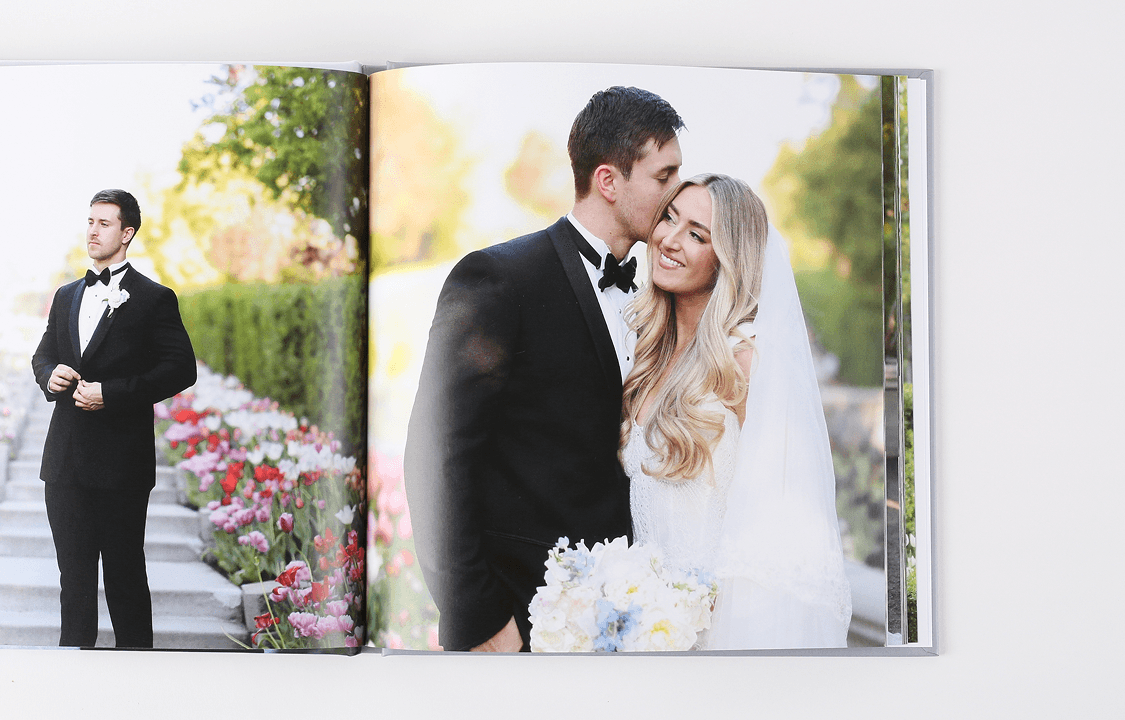 A wedding photo album open to two pages. Left: Groom in a tuxedo stands near a flower-lined path. Right: Bride and groom embrace, smiling with joy.